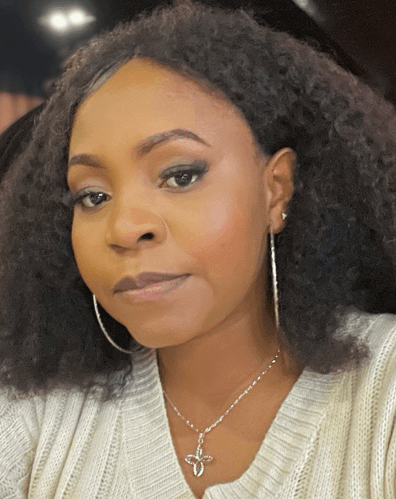 Woman with curly hair wearing hoop earrings and a cross necklace, dressed in a light sweater.