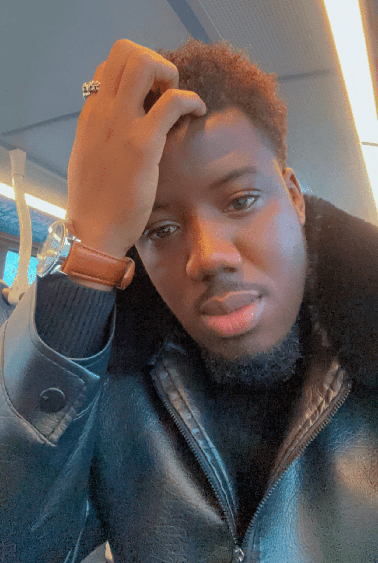 Person in a leather jacket sitting indoors, with hand on forehead and watch on wrist.