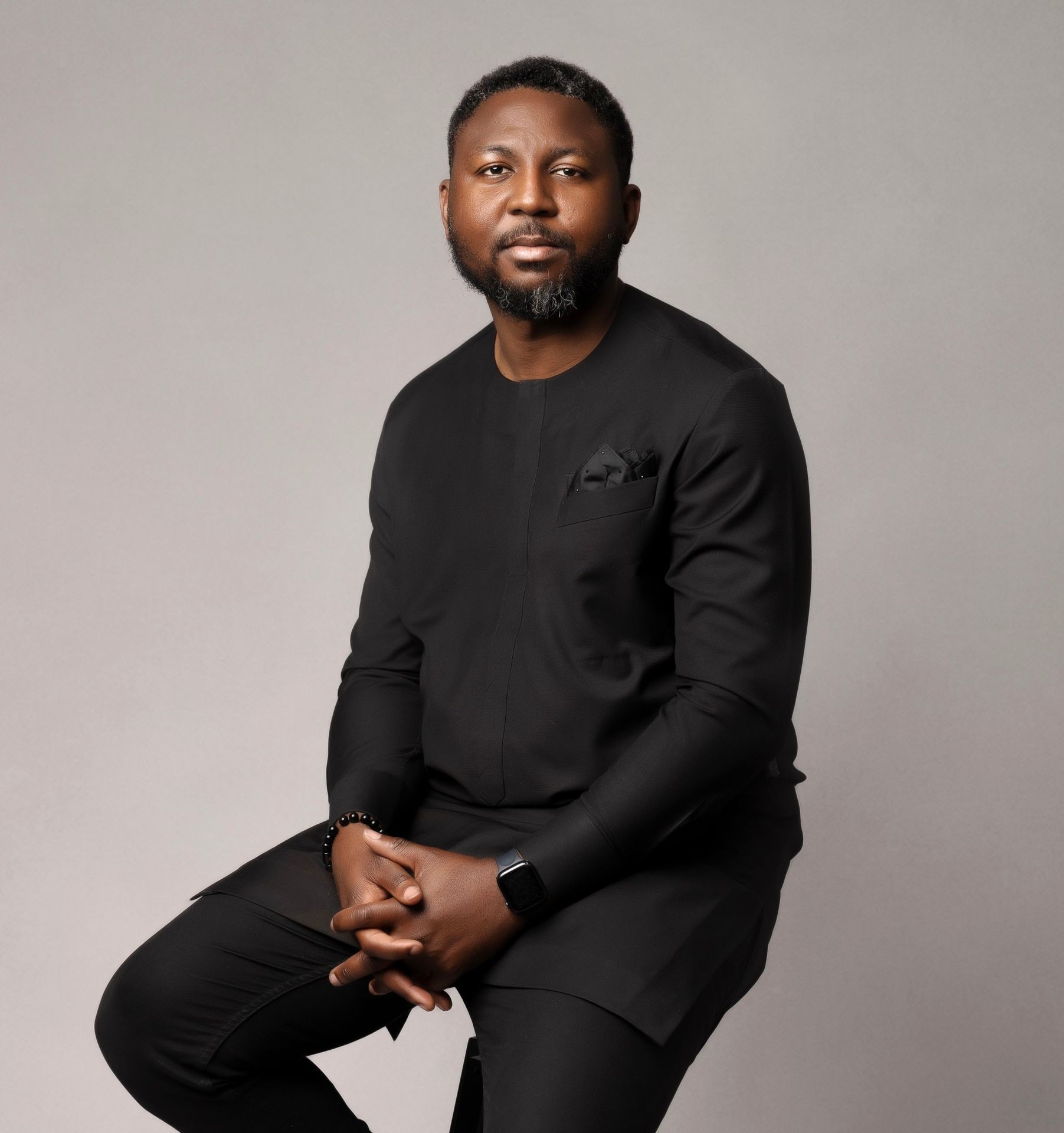 Man in a black outfit seated against a neutral background, wearing a watch and bracelet.
