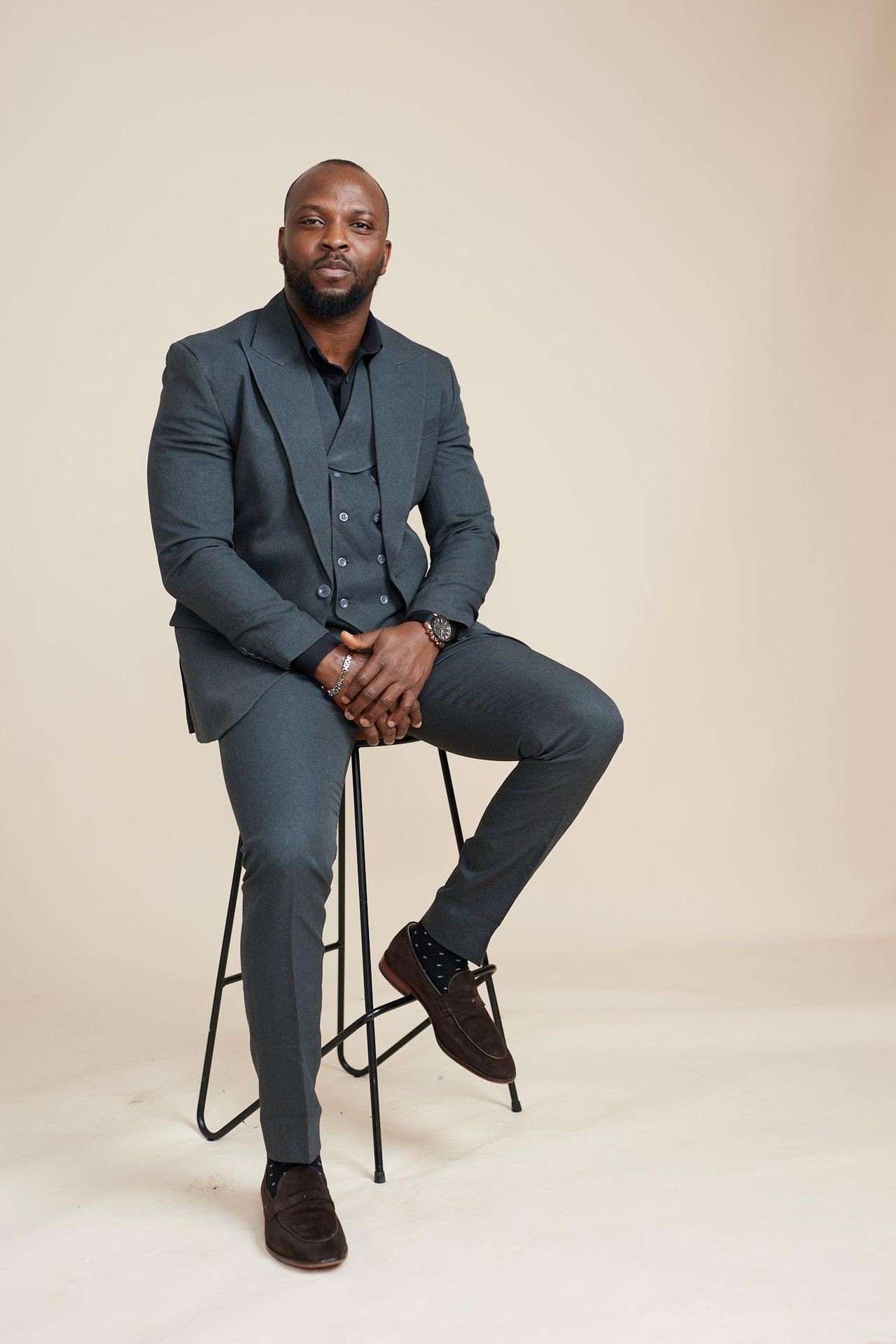Man in a dark grey suit sitting on a stool against a beige background.