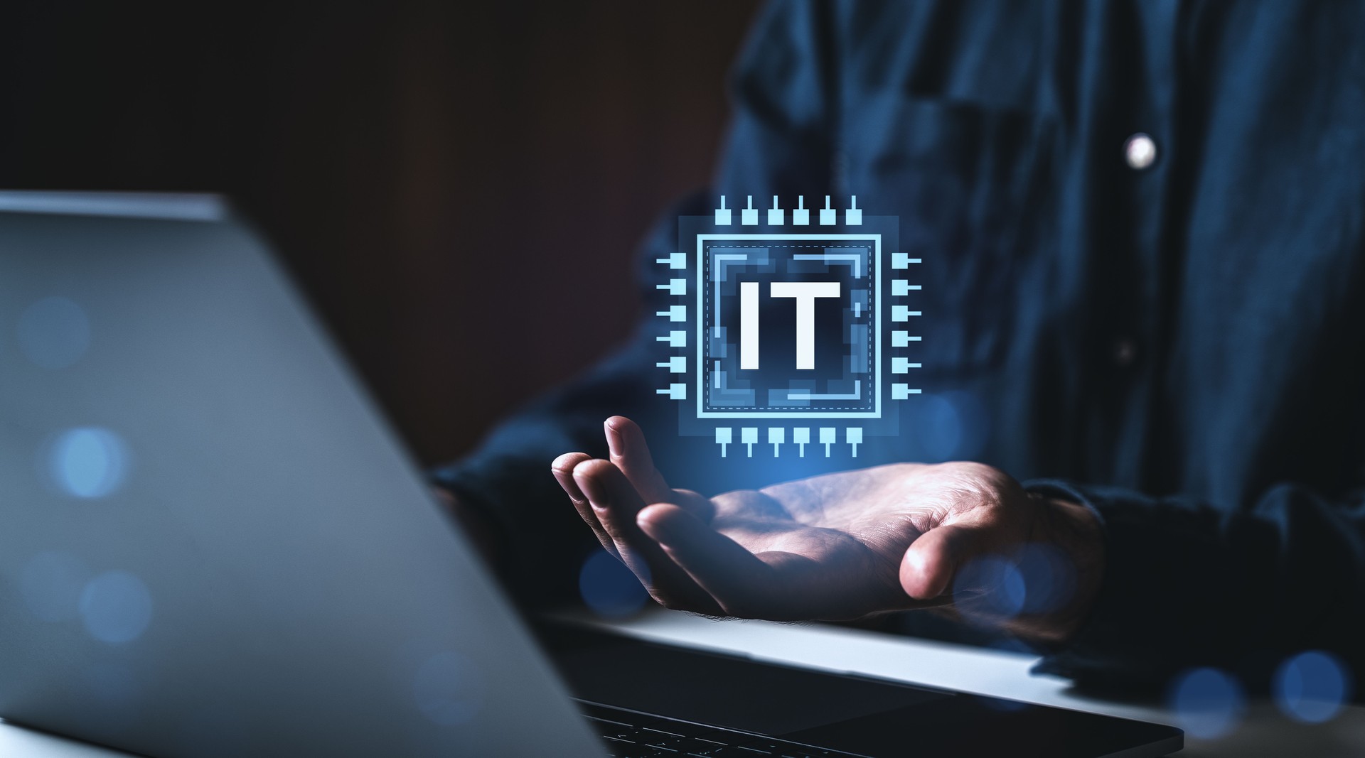 Information technology icon floating over open hand near laptop in dark room.